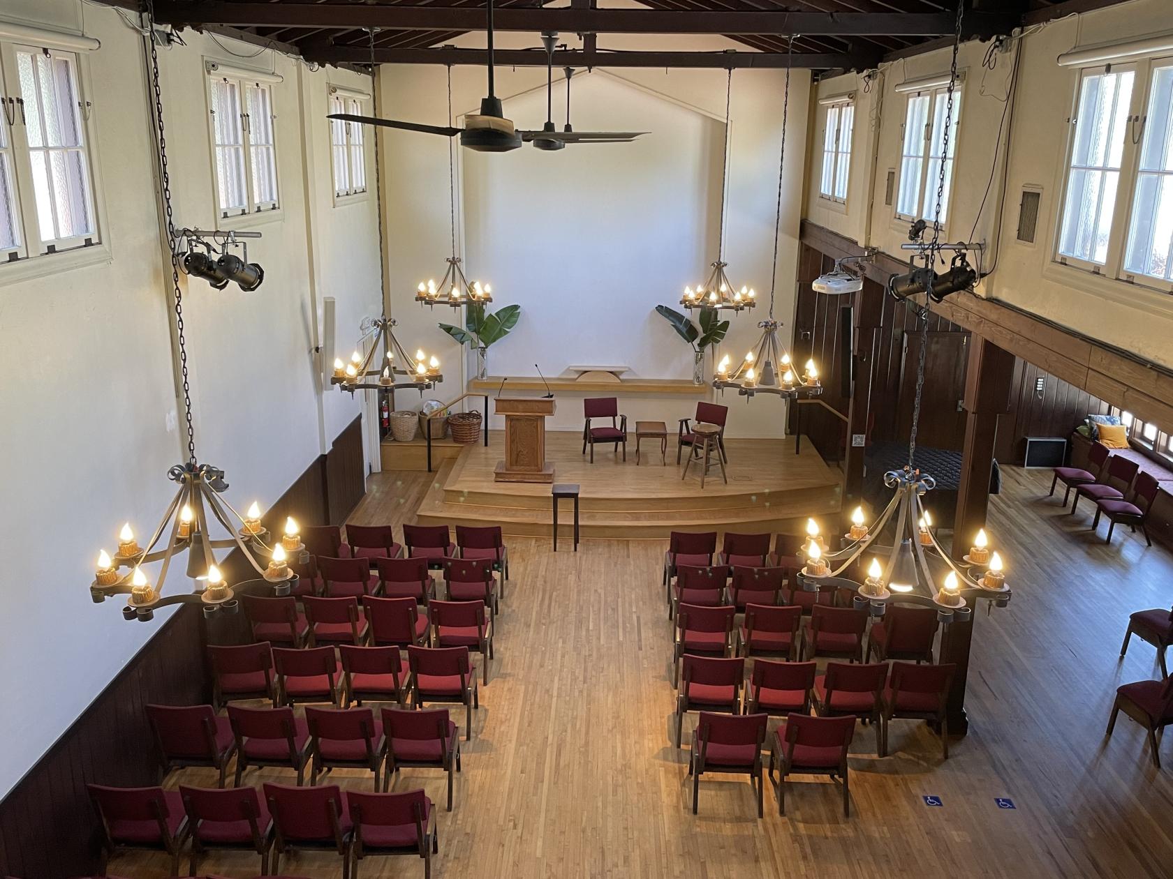 A church hall with red cushioned chairs, wooden flooring, chandeliers, and a raised stage at Unitarian Universalist Church of Studio City.
