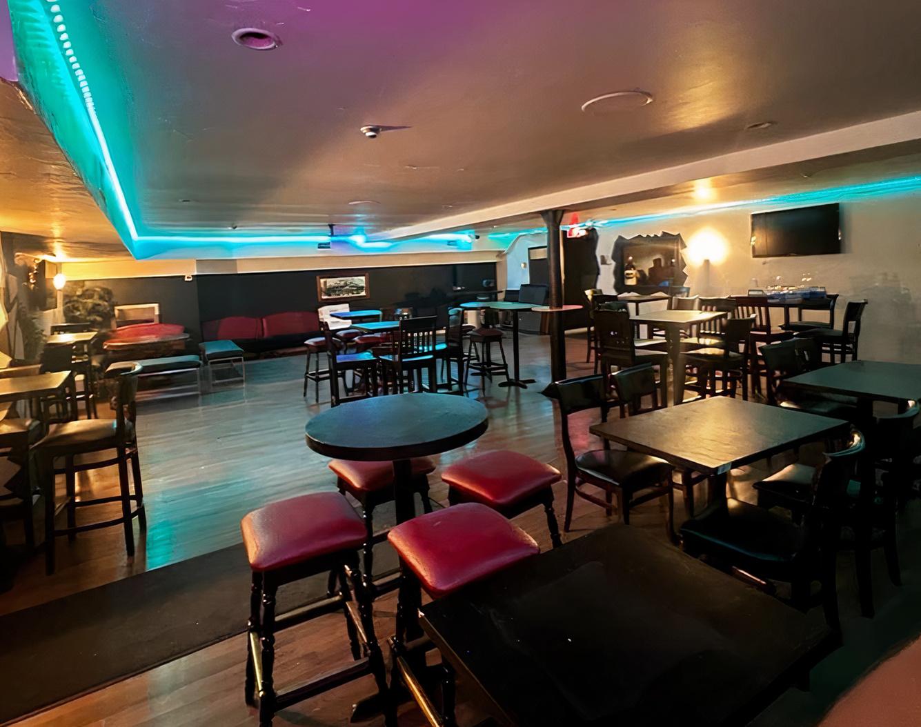 A basement event room with round and square tables, wood flooring, neon accent lighting, and wall-mounted TV at Fox and Fiddle - Danforth.