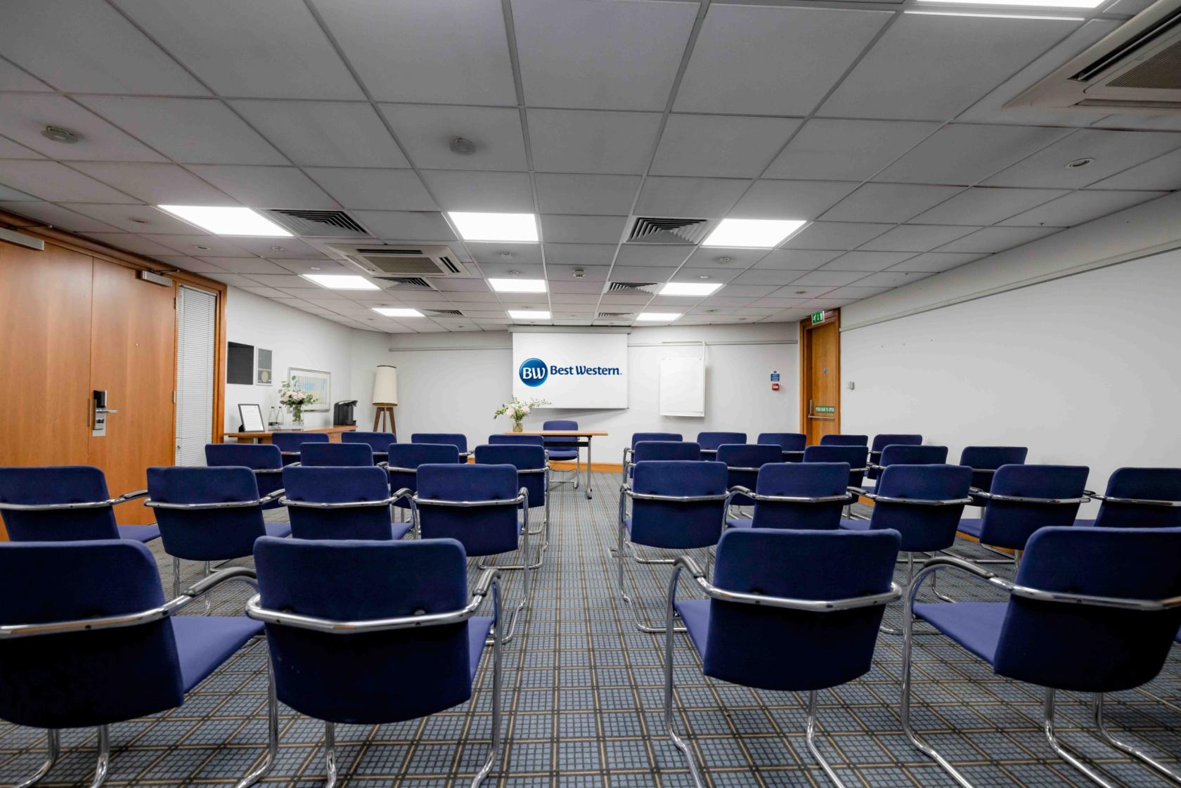 A hotel conference room with blue cushioned chairs, a projector screen, and grid-patterned carpet at Best Western Ariel London Heathrow.
