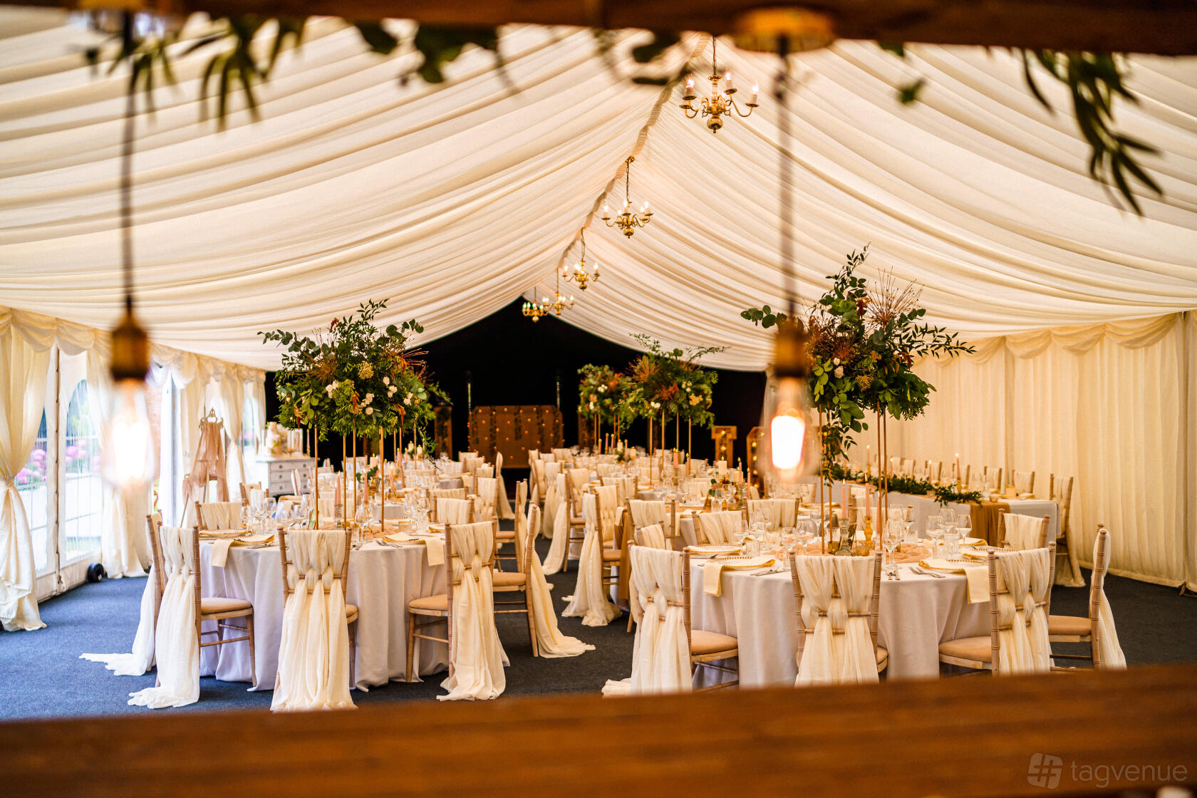 An outdoor event space with draped marquee ceiling, round tables, white linens, and tall floral centerpieces at Deene Park.