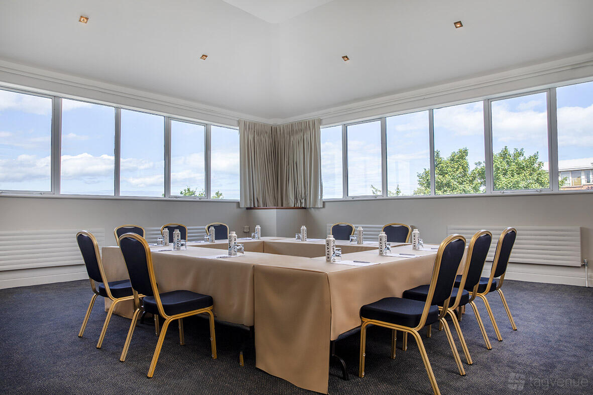 A meeting room with floor-to-ceiling windows, U-shaped tables with beige linens, and navy chairs at Grand Hotel.