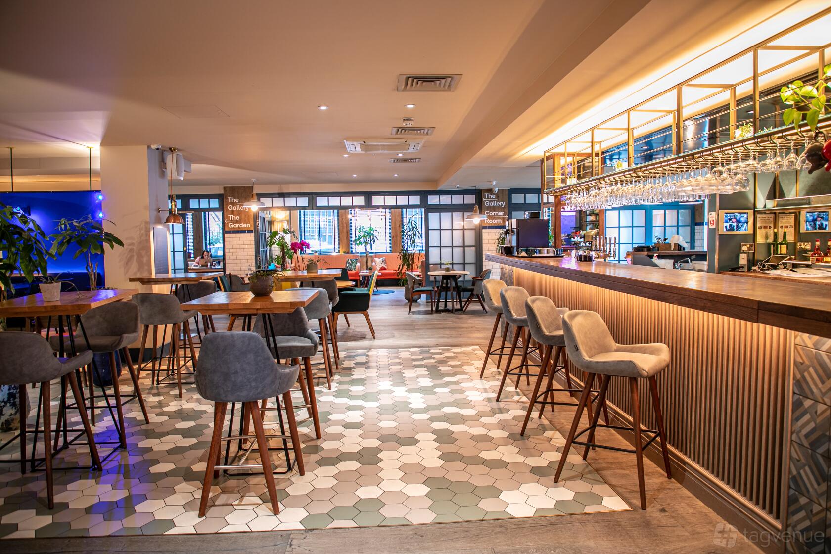 A restaurant in a hotel with a curved bar, pendant lighting, and gray upholstered bar stools at The Corner London City.