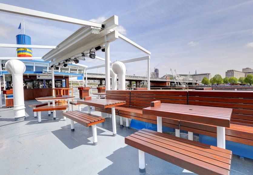An open-air boat deck with wooden benches, picnic tables, and a bar counter at The Tattershall Castle.