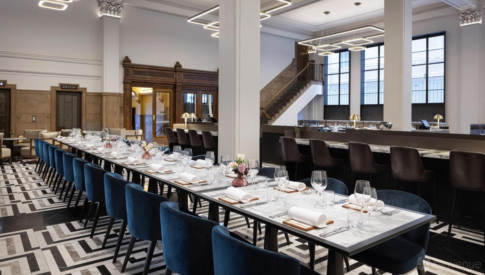 An event space in a hotel with long marble tables, velvet chairs, geometric patterned flooring, and large windows at Hilton Melbourne Little Queen Street.