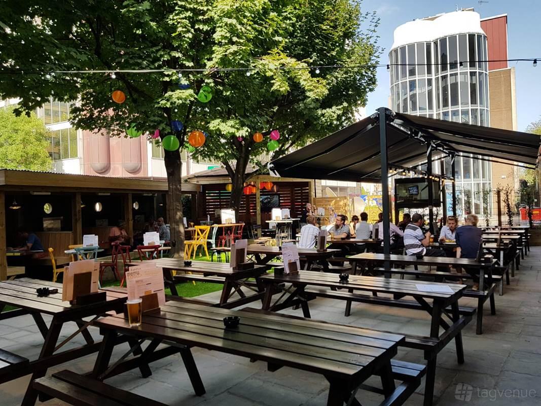 An outdoor pub garden with wooden picnic tables, colorful hanging lanterns, and mature trees at The Minories.