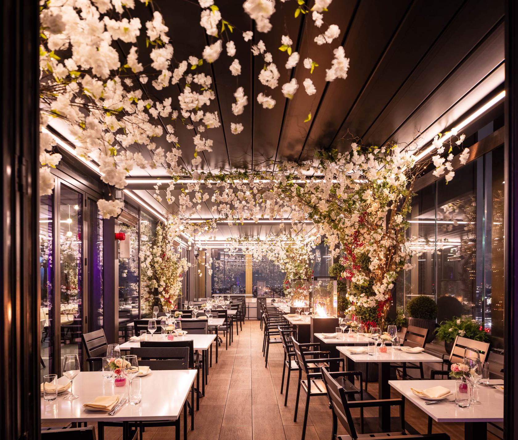 A rooftop terrace with floor-to-ceiling windows, floral ceiling installations, and set dining tables at STK Rooftop - Stratford.