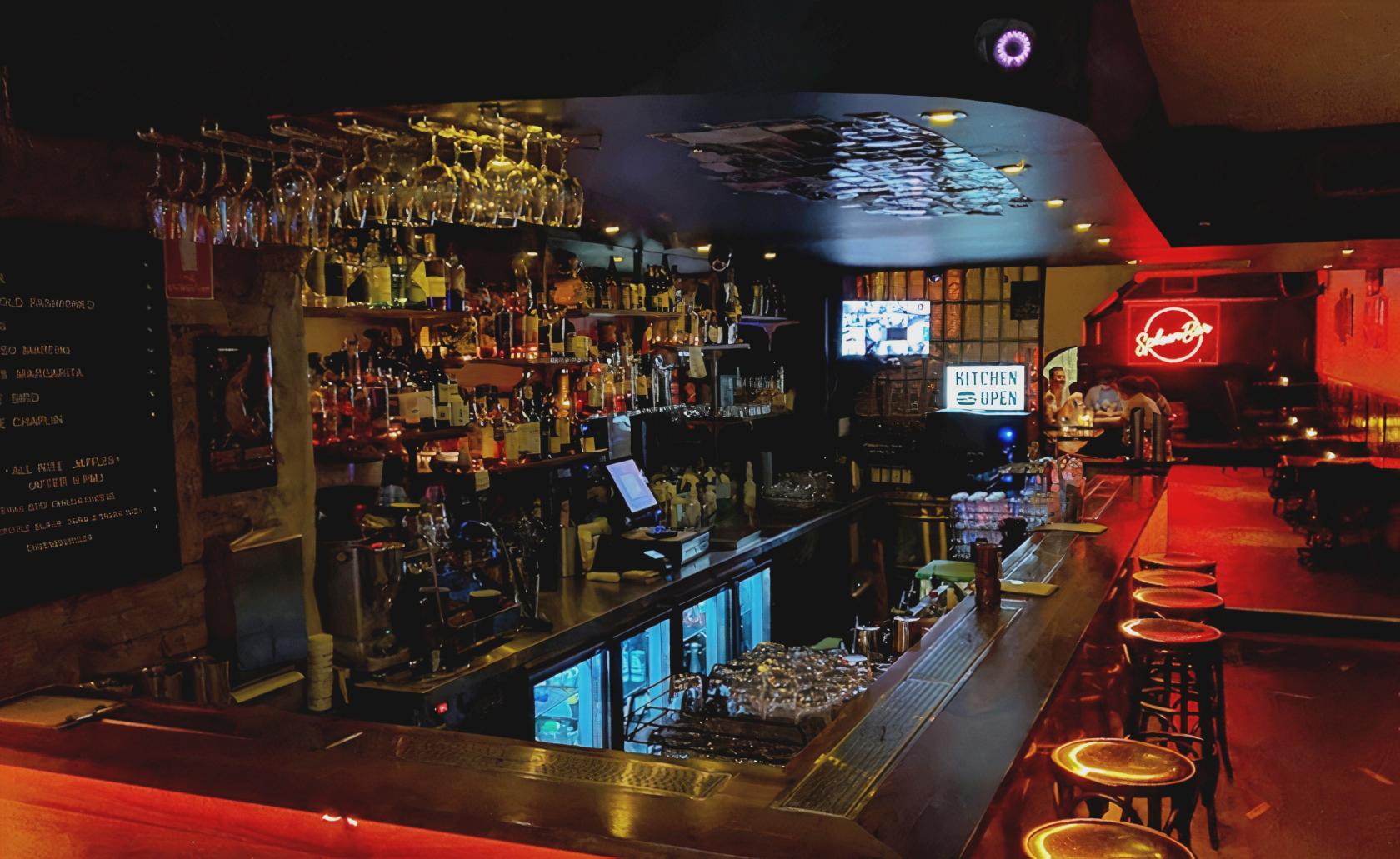 A bar with hanging glassware, a stocked liquor display, neon signage, and high stools at Spleen Bar.