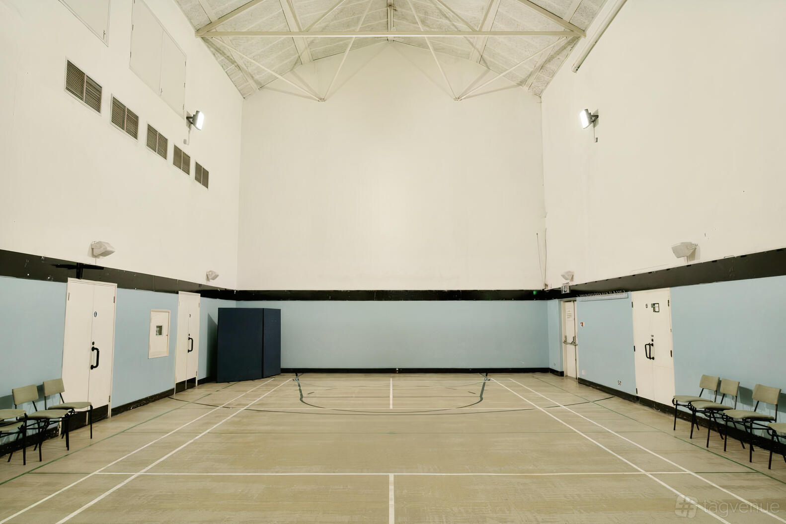 A community hall with high ceilings, blue and white walls, marked sports flooring, and rows of chairs at St Hilda's East.