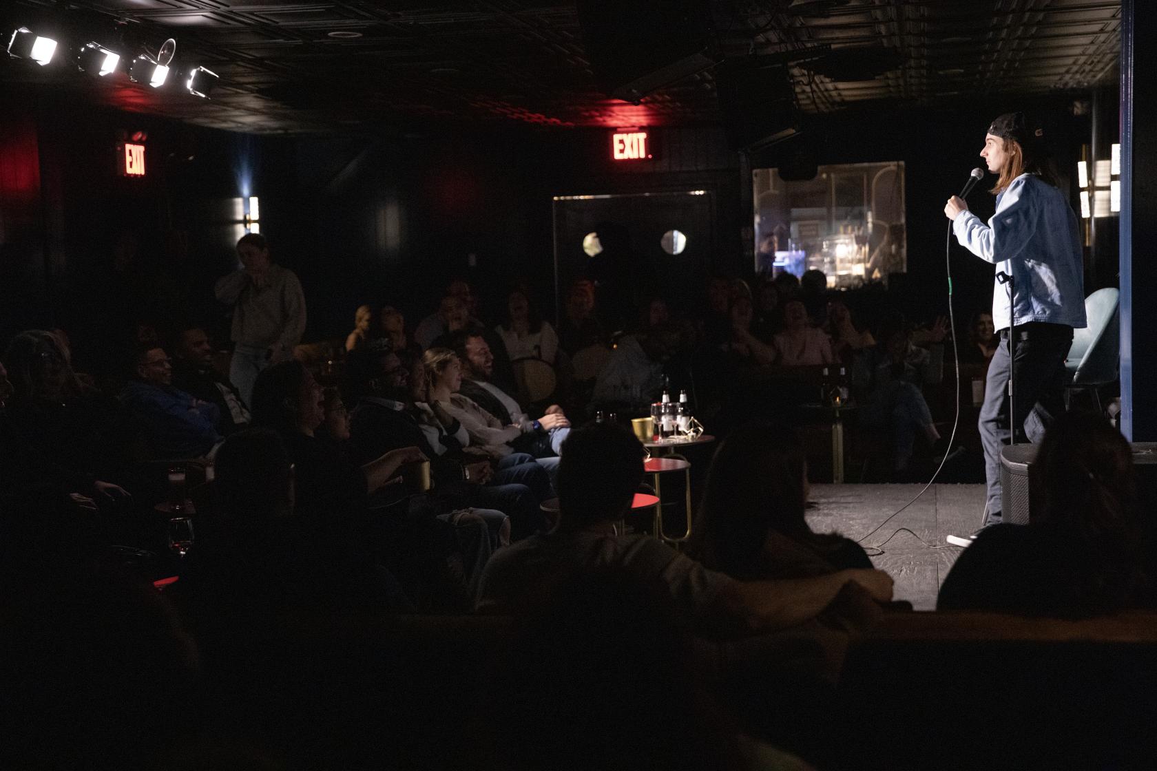 A dimly lit bar with a stage, live stand-up performance, and seated audience at Rodney’s.