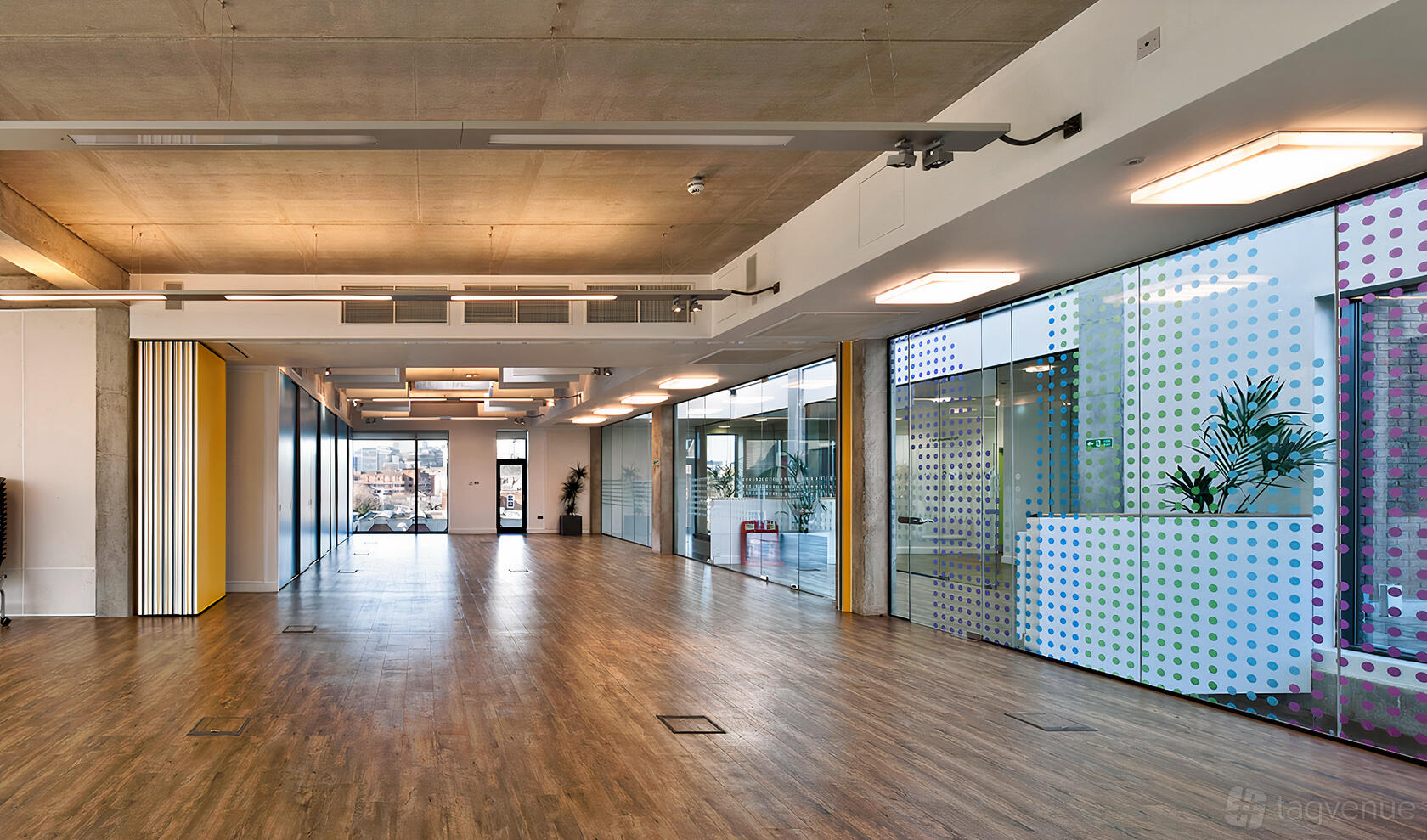 A hall with floor-to-ceiling glass windows, wooden floors, and exposed concrete ceiling at Osmani Centre.