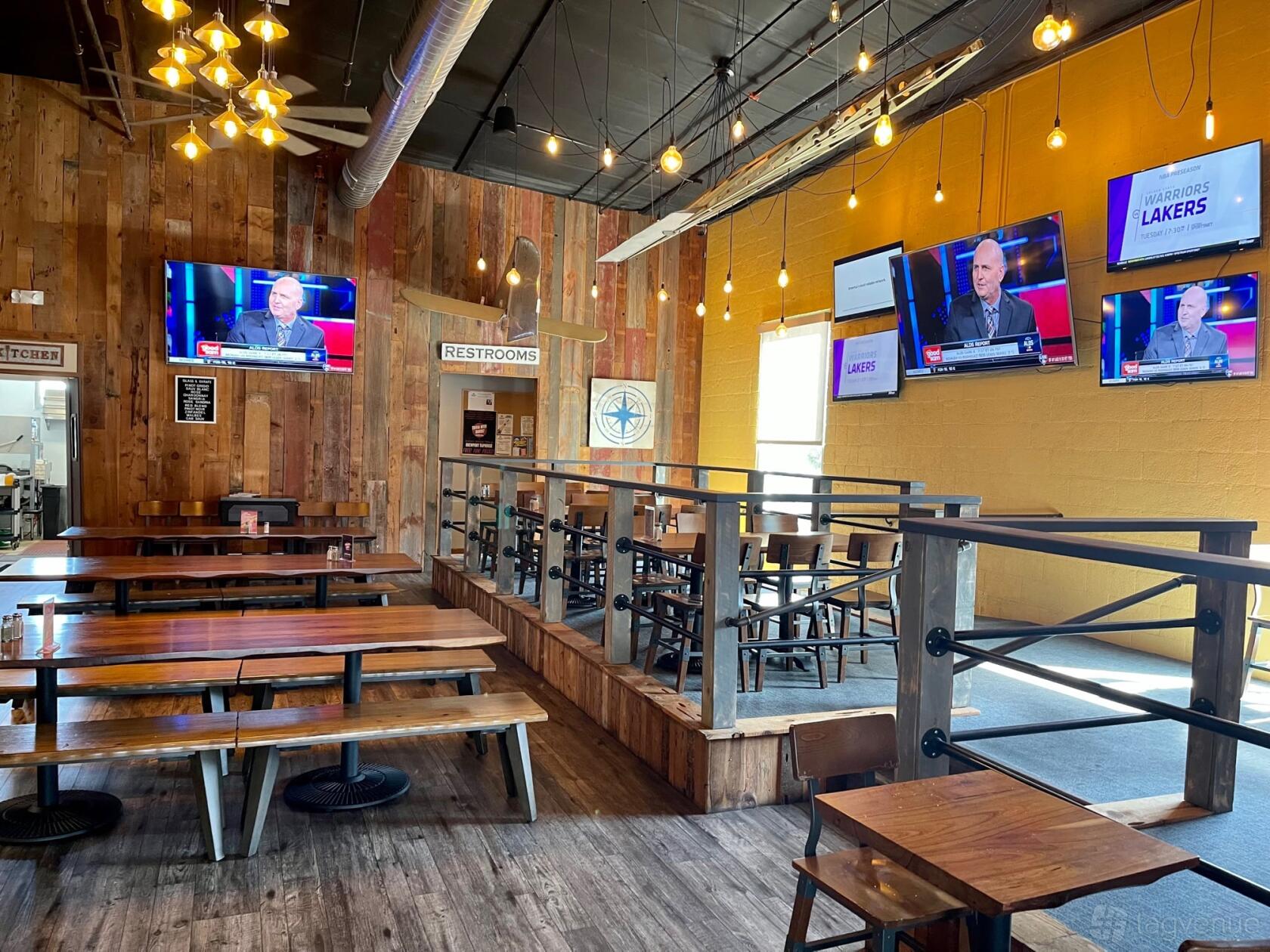A bar in a restaurant with wooden communal tables, TV screens, and exposed bulb lighting at Brewport Tap House.