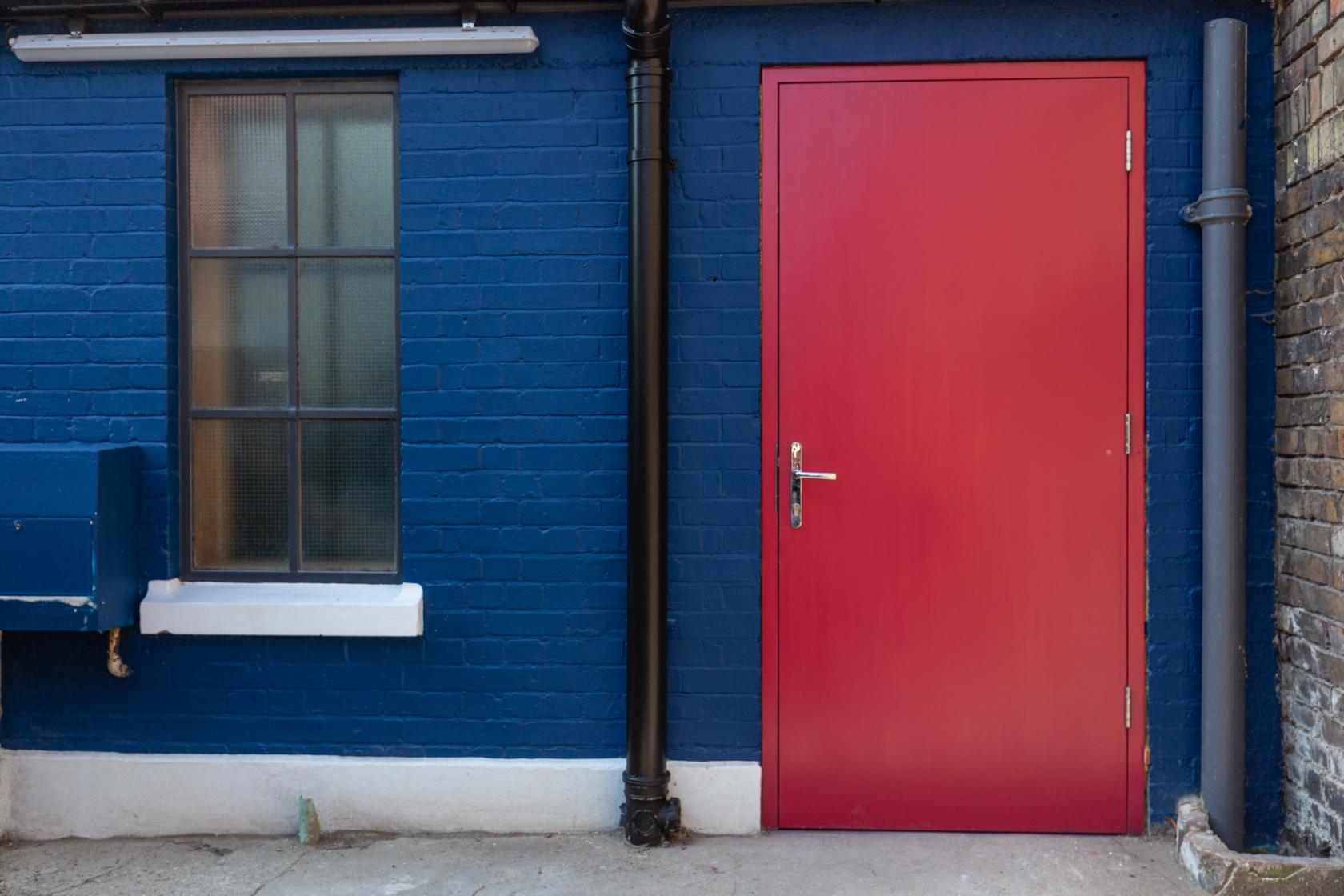 A vibrant blue brick exterior with a red door and industrial window at Fomo London Studio.