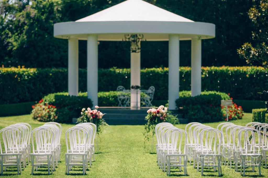 An outdoor event space with a white pavilion, white chairs arranged on the lawn, and floral arrangements at Miramare Gardens.