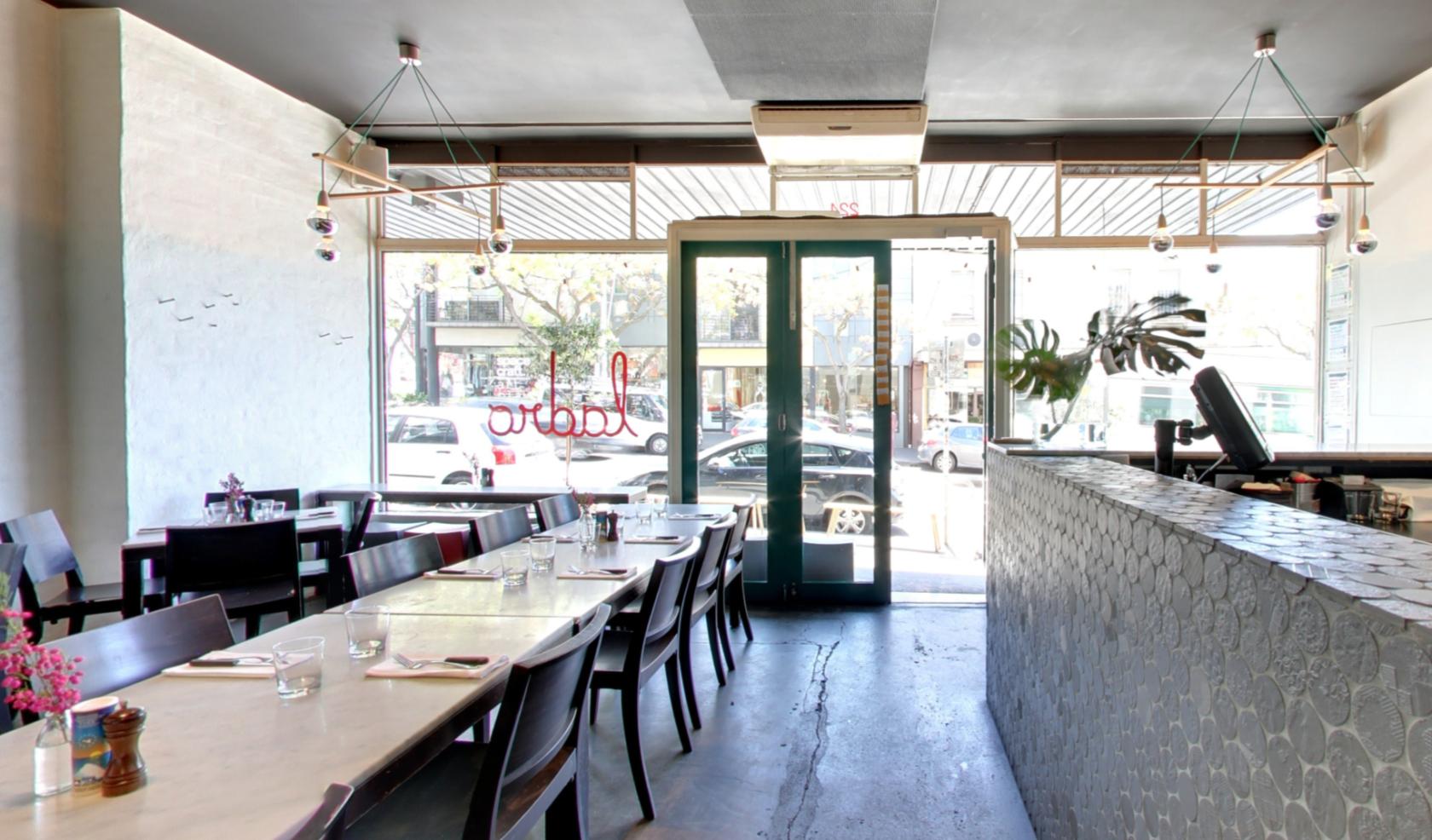 A dining room with long marble tables, dark wooden chairs, and large floor-to-ceiling windows at Ladro Restaurant.