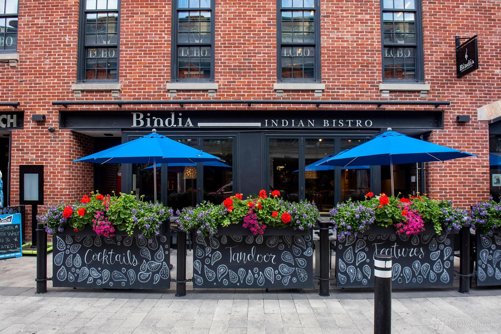 A restaurant patio with blue umbrellas, floral planter boxes, and brick facade at Bindia Indian Bistro.