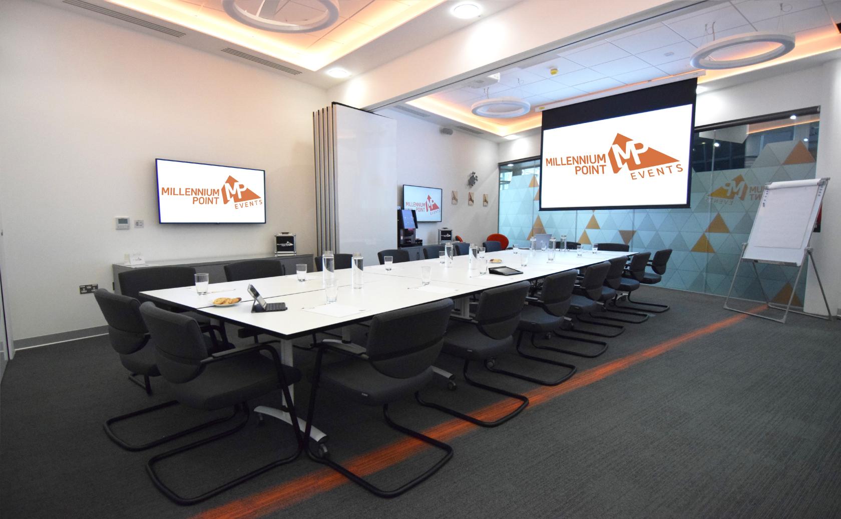 A conference centre meeting room with a long table, black chairs, presentation screens, and glass walls at Millennium Point.