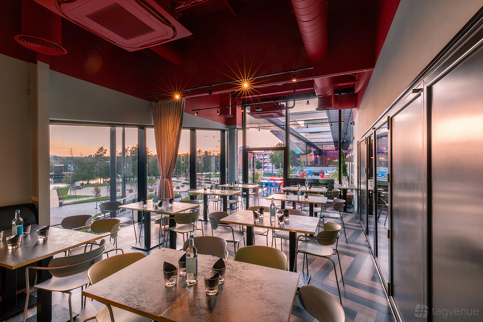 A dining room with floor-to-ceiling windows, exposed red ceiling pipes, and set tables at Puttshack Lakeside.