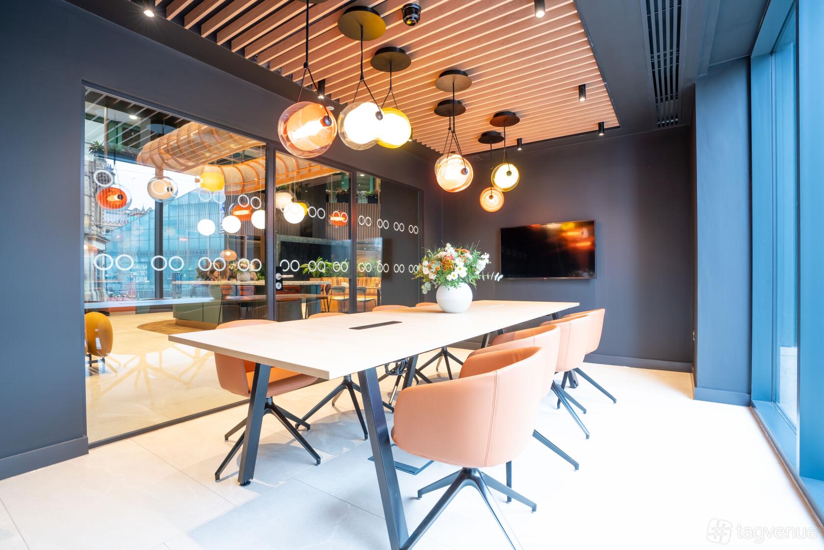 A meeting room with pendant lighting, a long wooden table, orange chairs, and a wall-mounted screen at Orega Balloon Street.