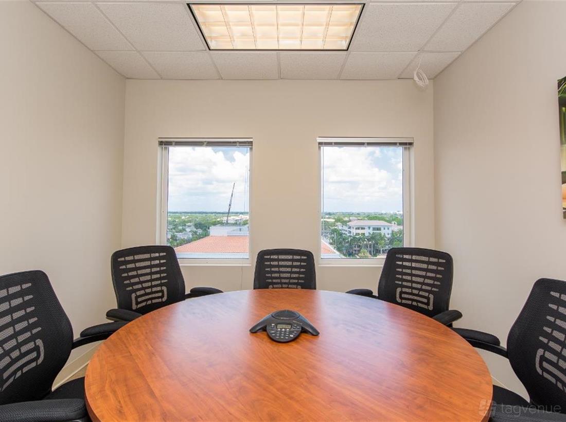 A meeting centre with a round wooden table, black office chairs, and two large windows at ZEN Offices Boca Raton.