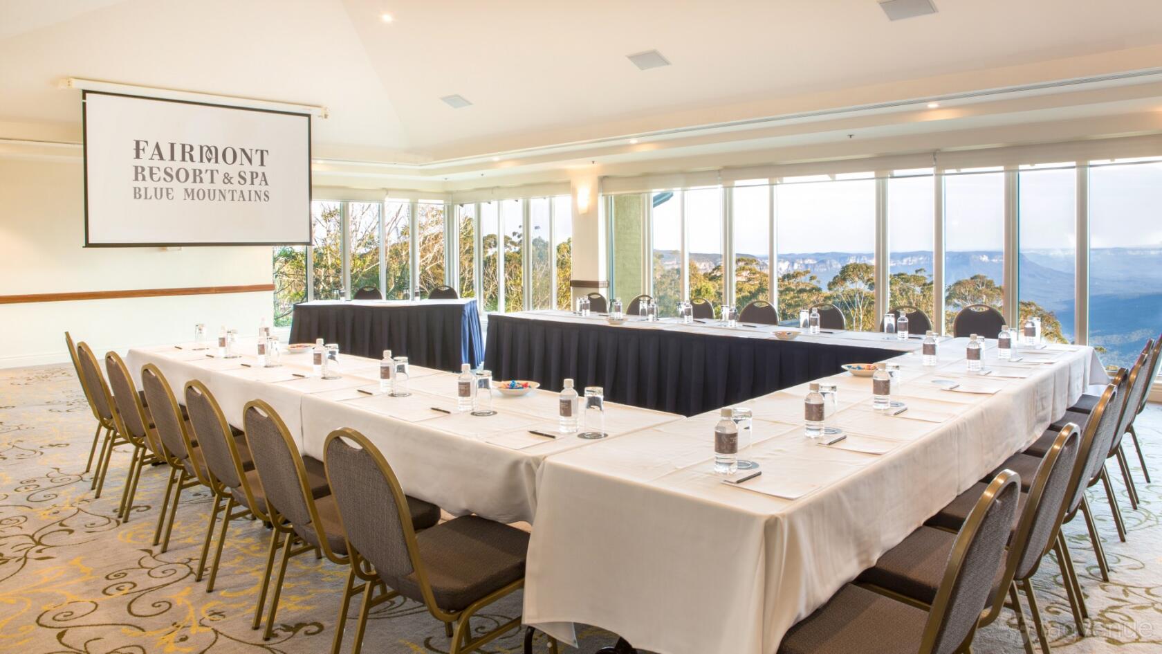 A meeting room with large windows, U-shaped tables, water glasses, and a projection screen at Fairmont Resort Blue Mountains.