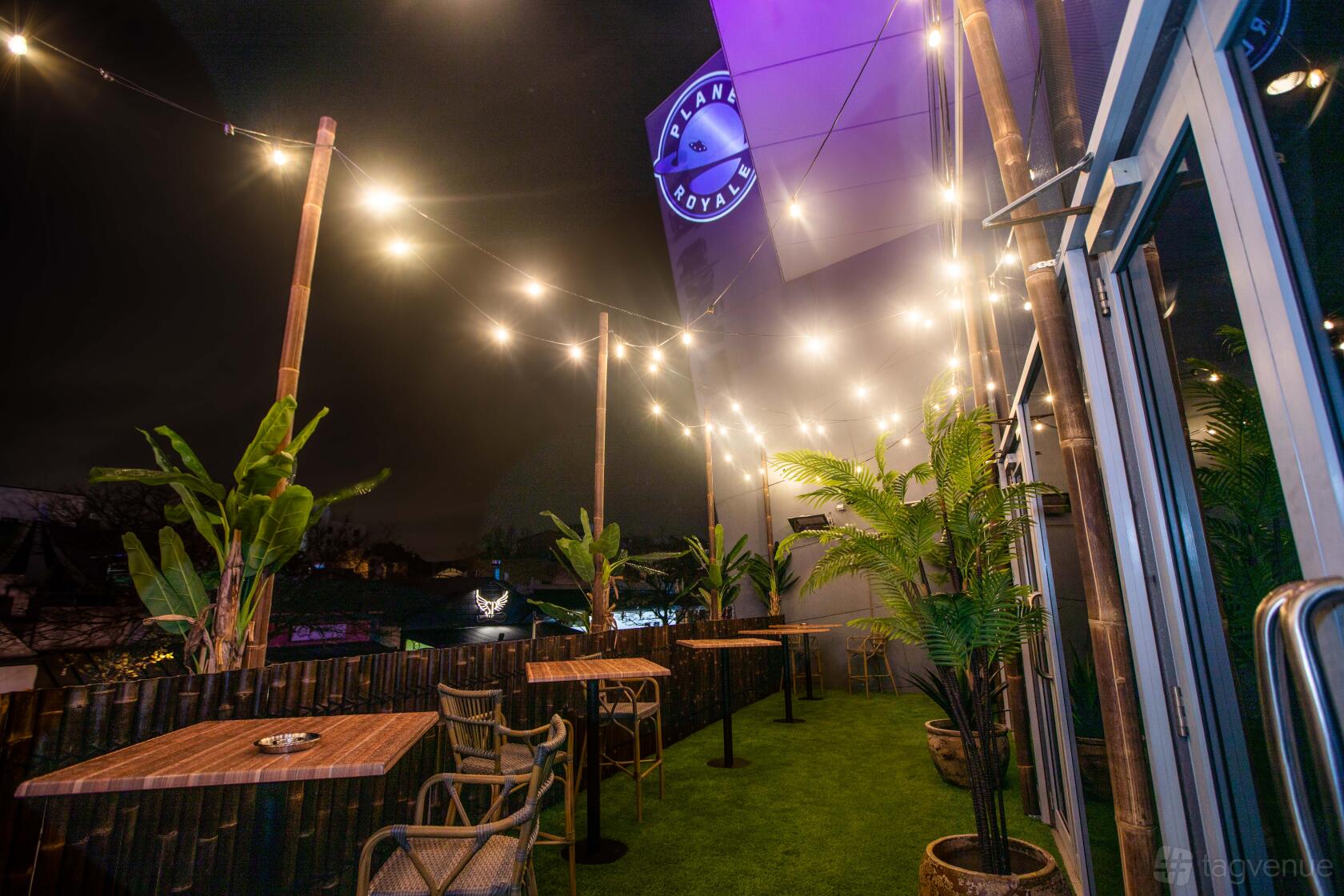 An outdoor bar with string lights, potted palms, and wooden tables on artificial grass at Planet Royale.