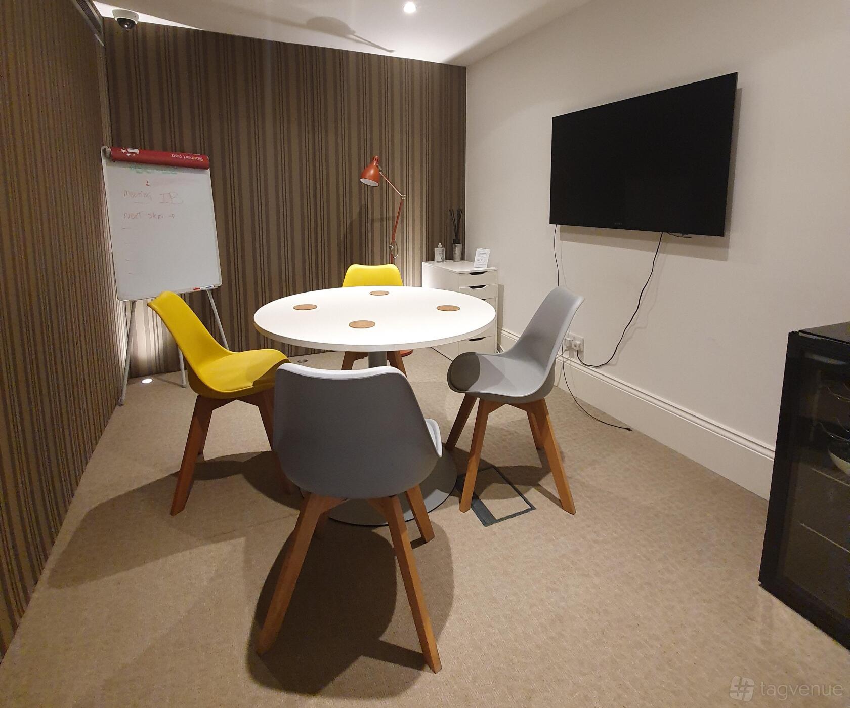 A meeting room with a round white table, four colorful chairs, a wall-mounted TV, and a flip chart at IdeaSpace London.
