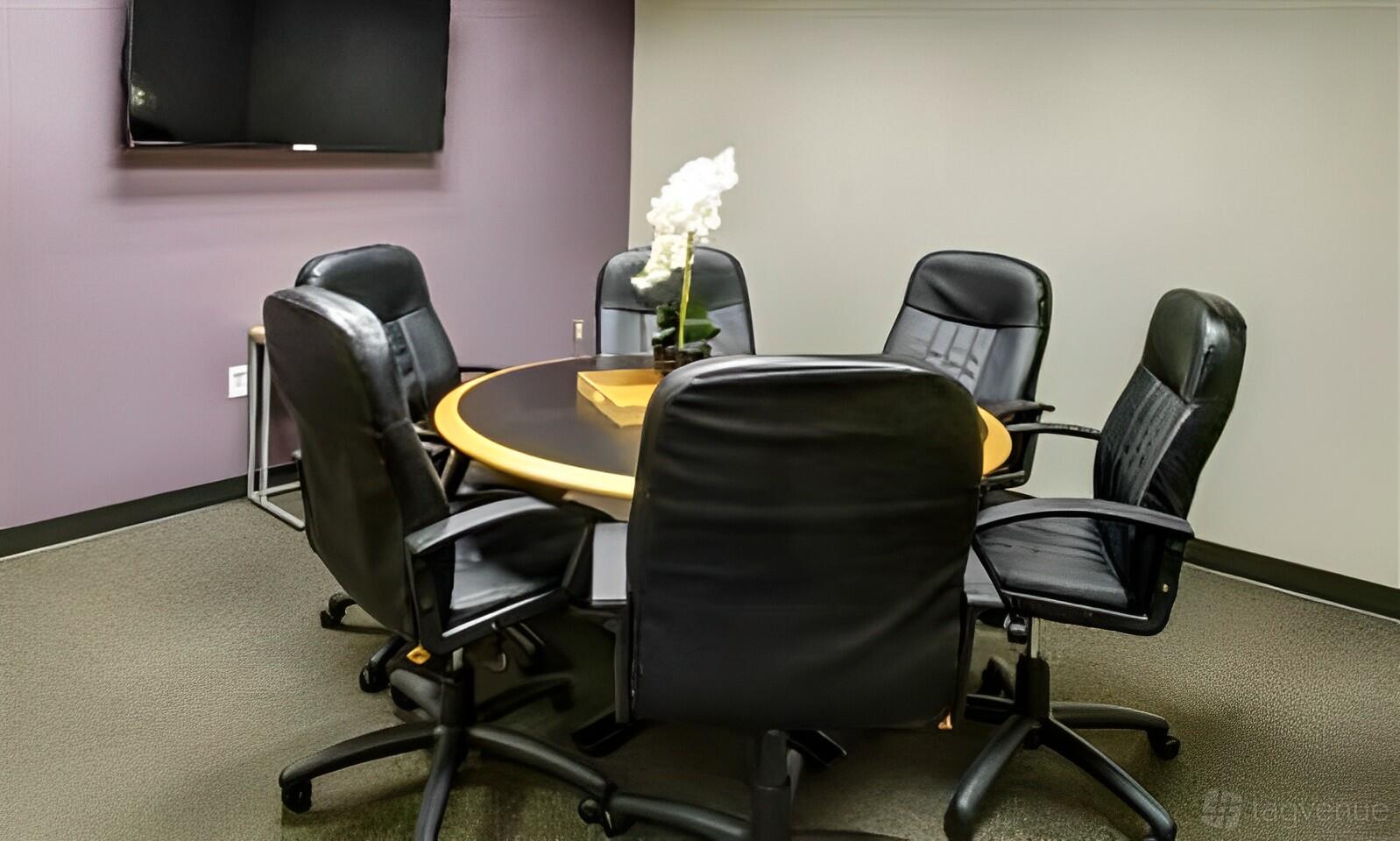 A meeting room with a round wooden table, six black leather chairs, wall-mounted TV, and a white orchid centerpiece at Intelligent Office in Rockville, MD.