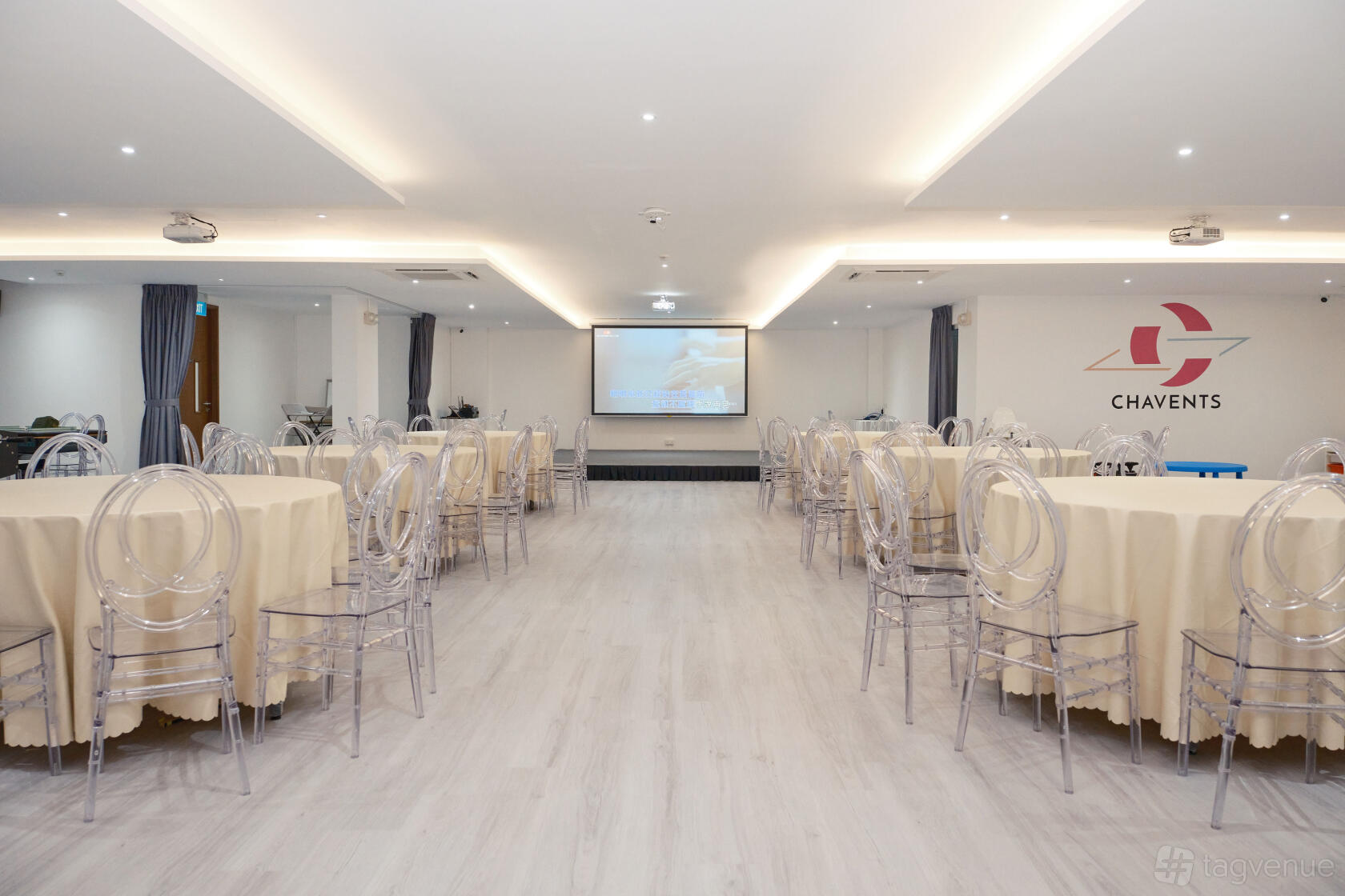 An event venue with round tables covered in beige linens, clear chairs, and a large projector screen at Chavents - Events For All.