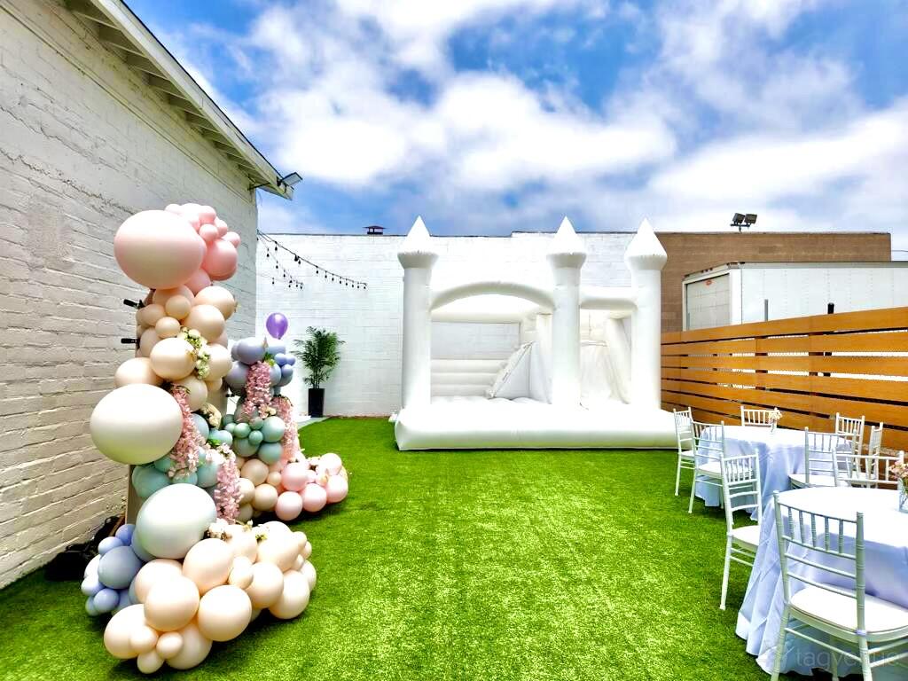 An event venue with artificial grass, a white bounce house, pastel balloon decorations, and round tables at KUE Event Space.