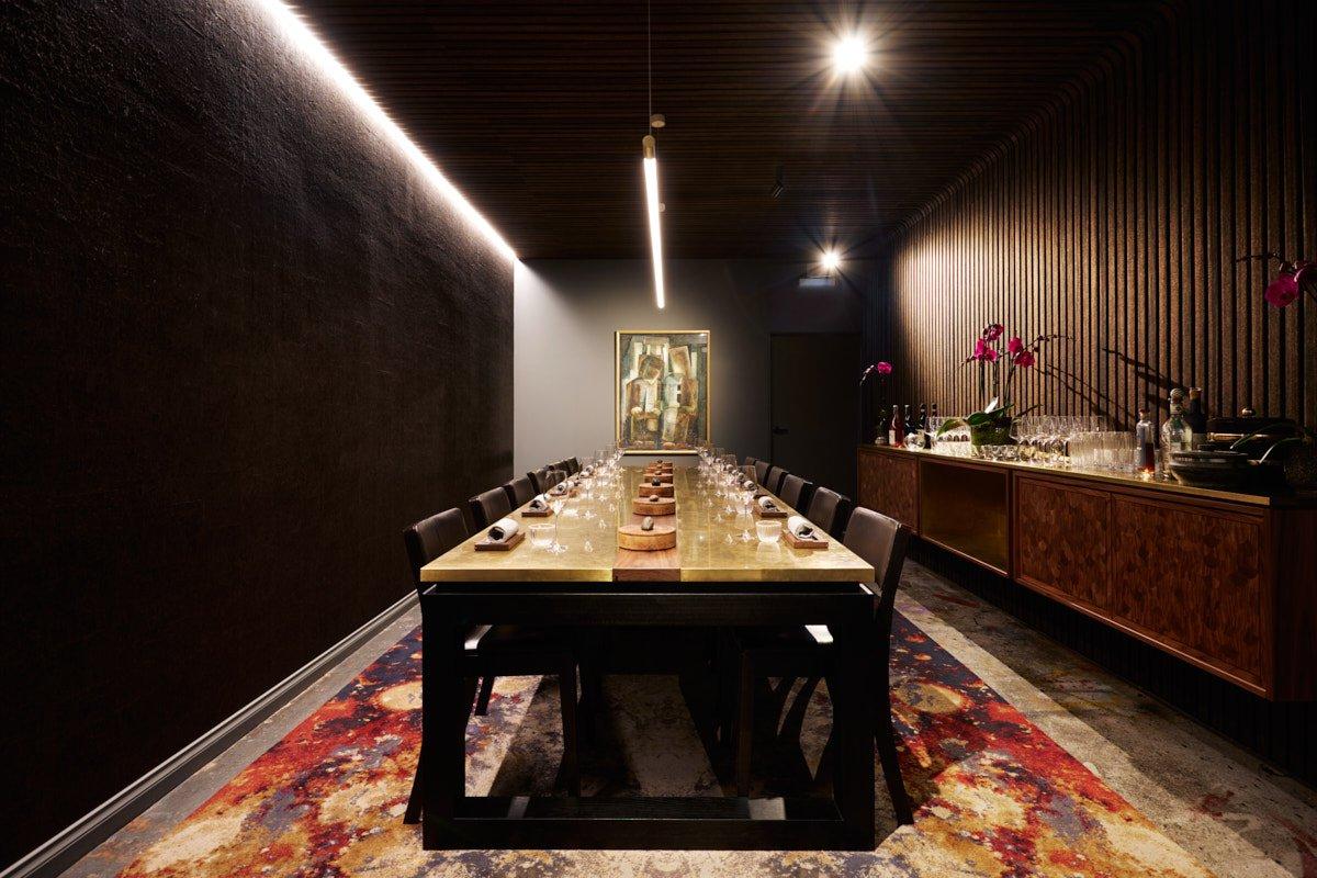 A dining room with a long brass table, dark walls, patterned rug, and a sideboard with orchids at I D E S.