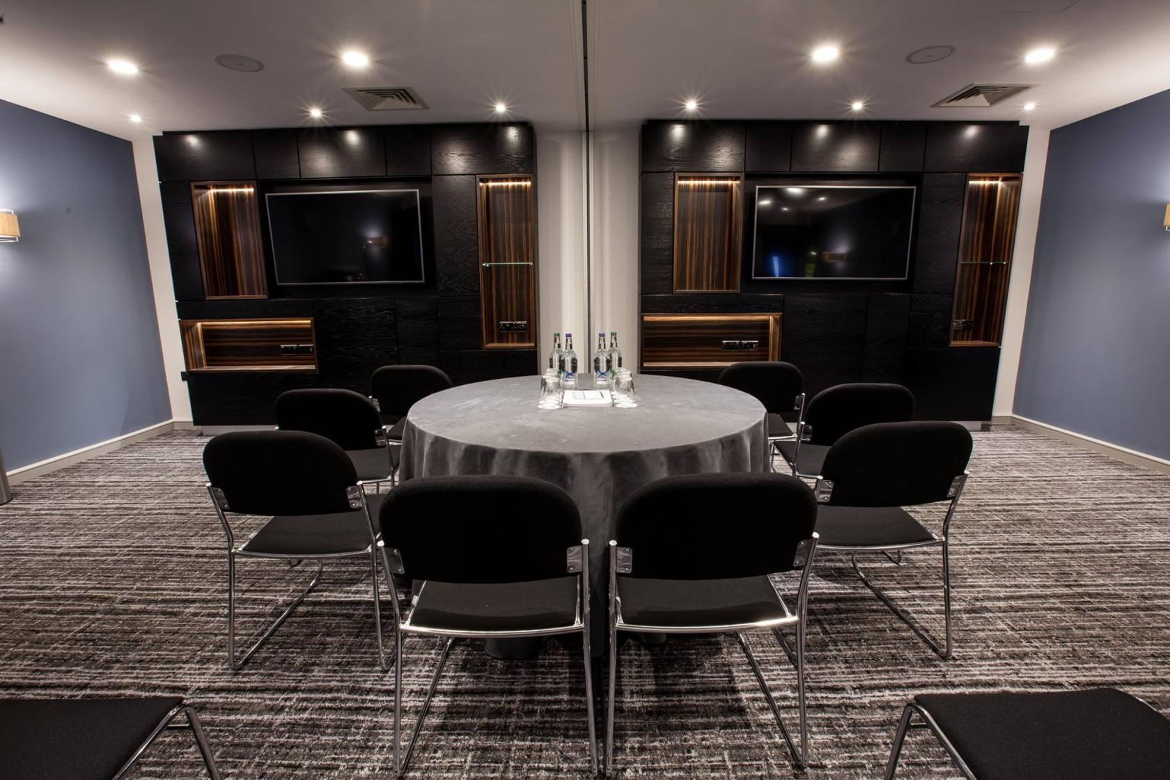 A hotel meeting room with a round table, black chairs, wall-mounted screens, and gray carpeting at The BCEC.