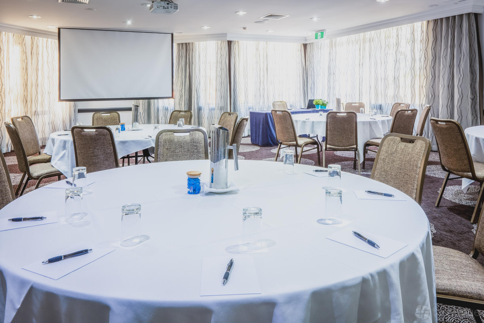 A hotel conference room with round tables set for meetings, notepads, pens, and a projector screen at View Brisbane.