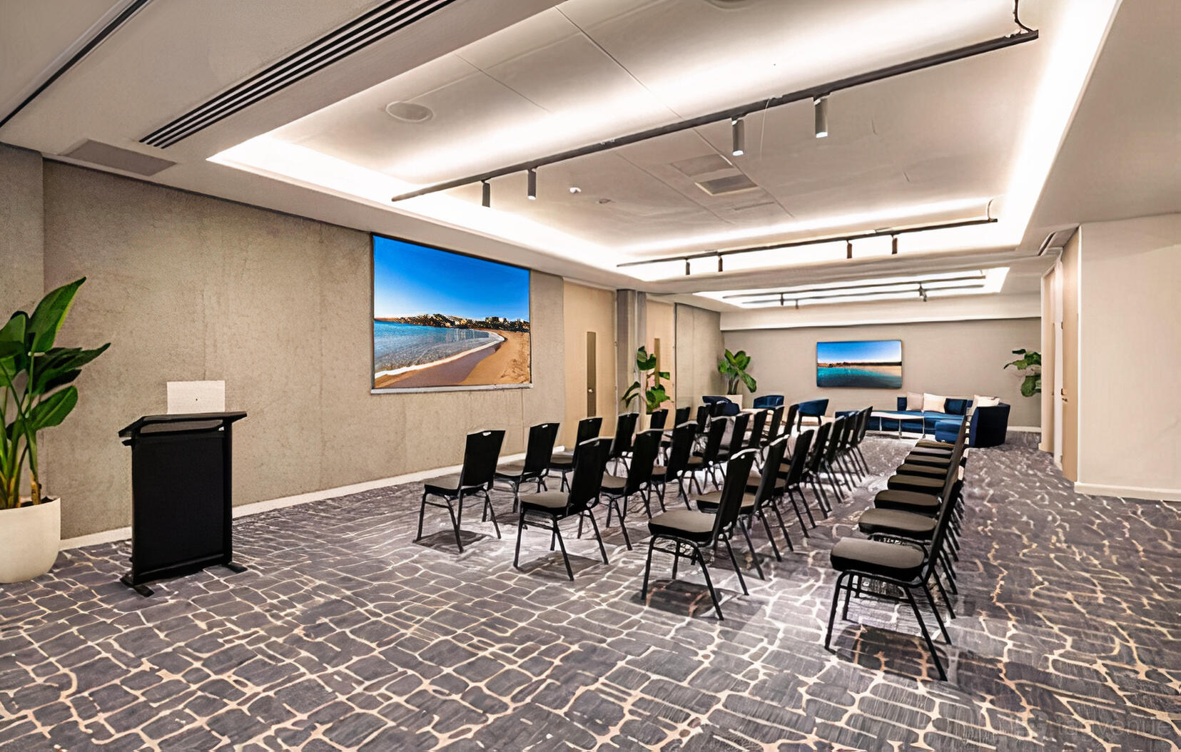 An auditorium with rows of black chairs, a podium, large wall screens, and potted plants at Crowne Plaza Sydney Coogee Beach.