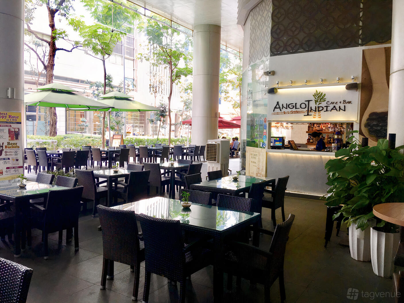 An outdoor cafe patio with wicker chairs, glass-topped tables, and green umbrellas at Anglo Indian Cafe & Bar - Shenton Way.