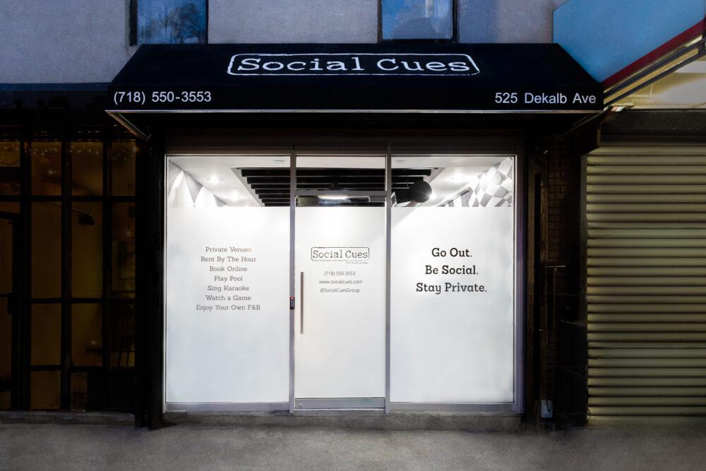 A storefront with white frosted glass windows, black awning, and bold signage at Social Cues - Surreal.