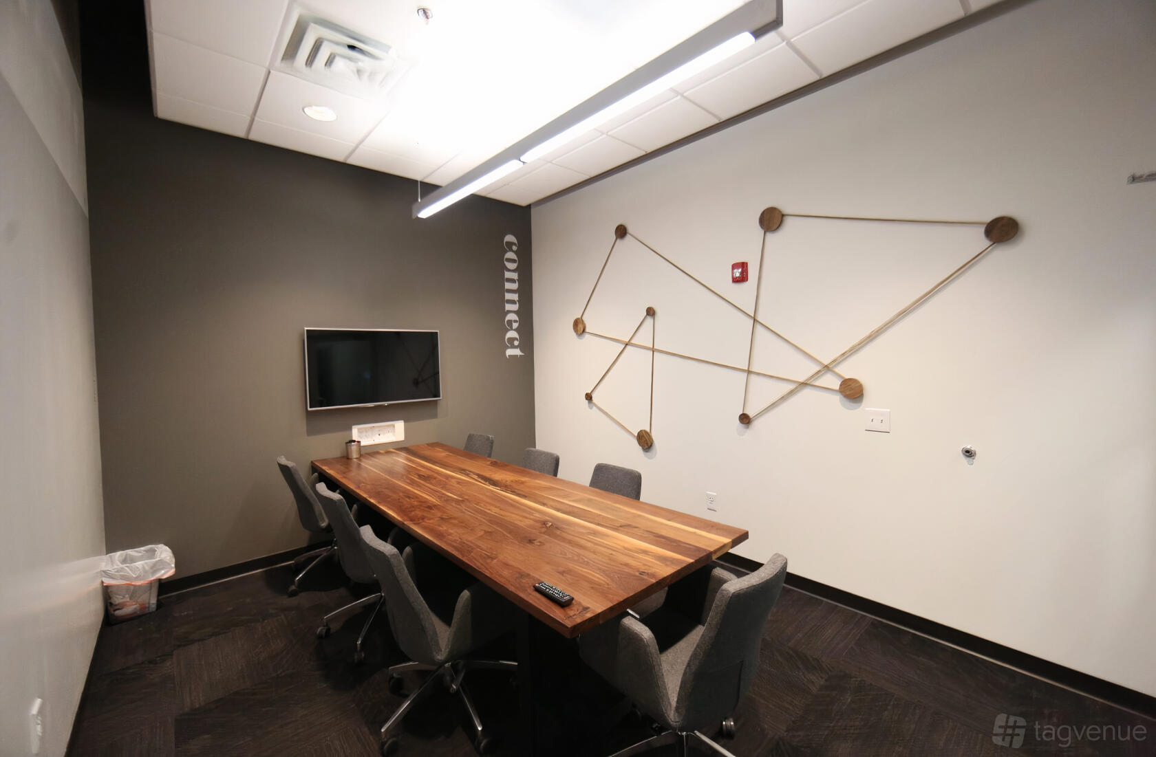 A meeting room with a long wooden table, six gray chairs, wall art with geometric patterns, and a wall-mounted TV at ROAM GALLERIA.