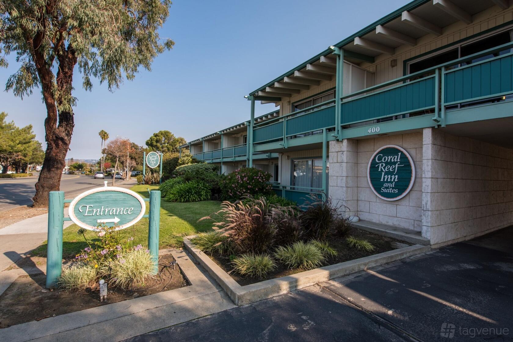 A hotel exterior with green railings, stone walls, and landscaped greenery at Coral Reef Inn and Suites.