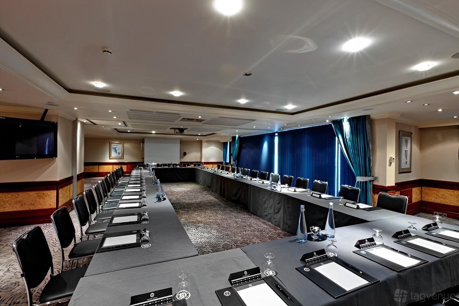 A hotel meeting room with U-shaped tables, black chairs, notepads, and blue curtains at Leonardo Royal London City.