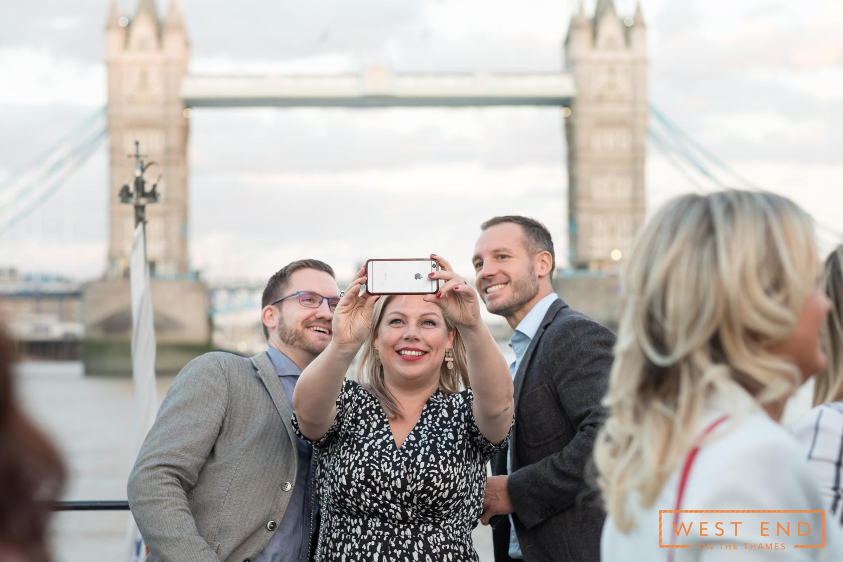 A boat event space with guests taking selfies on deck and views of Tower Bridge at West End on the Thames.