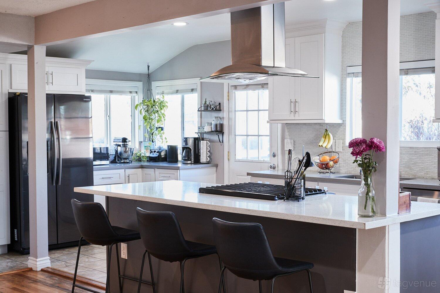 A house kitchen with a central island, stovetop, black barstools, stainless steel appliances, and bright windows at Lombard.