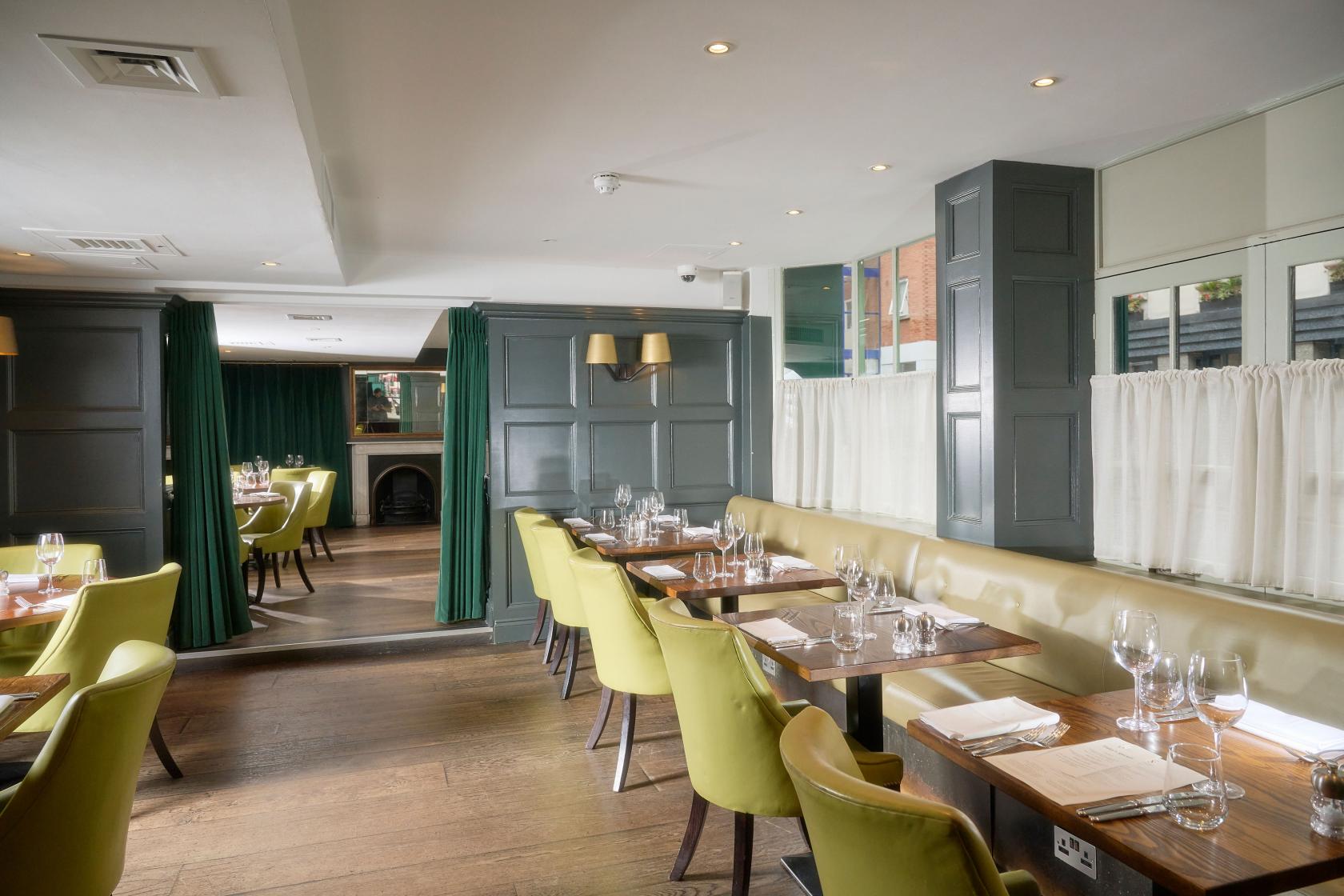 A pub with wood flooring, sage green chairs, and large windows with curtains at Chiswell Street Dining Rooms.