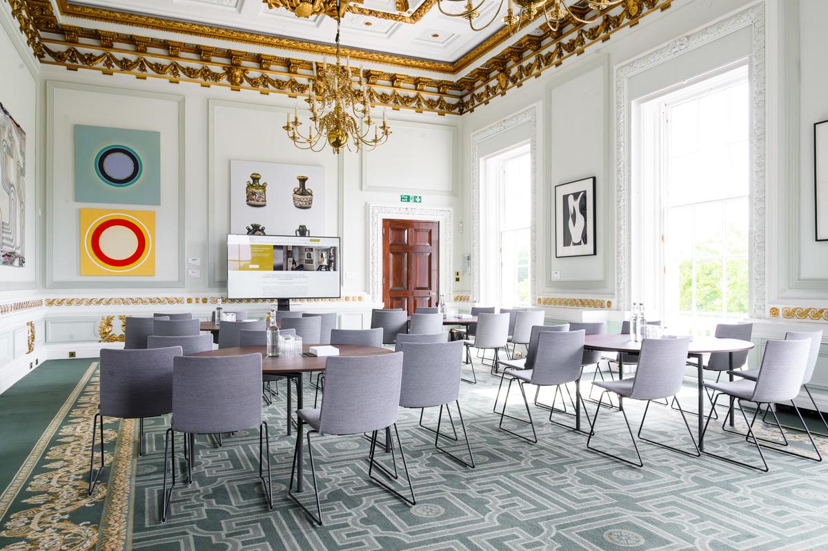 A function room with ornate gold ceiling moldings, large windows, abstract art, and grey chairs at {10-11} Carlton House Terrace.