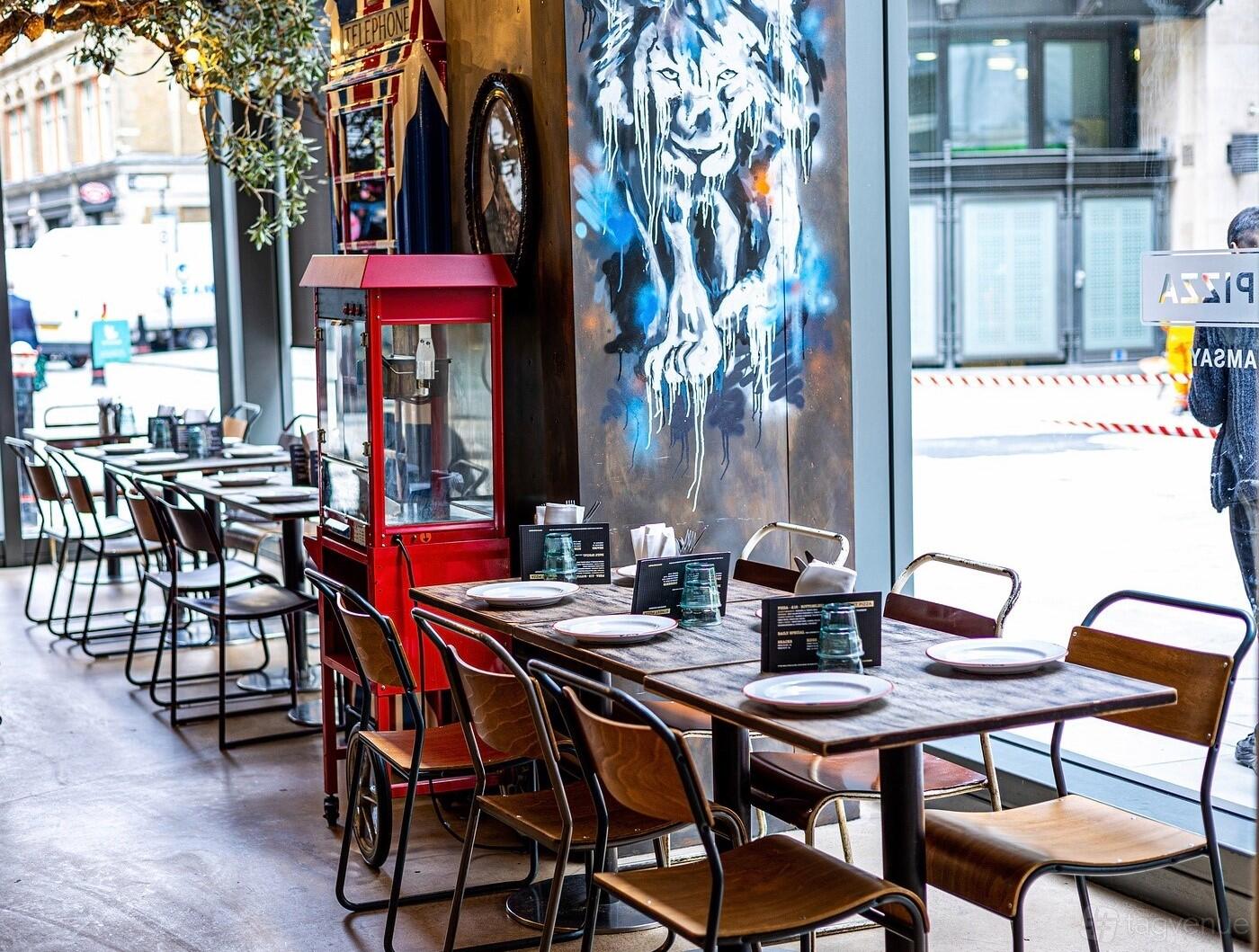 A restaurant dining area with wooden tables, metal chairs, floor-to-ceiling windows, and a mural at Bread Street Kitchen, St Paul's.