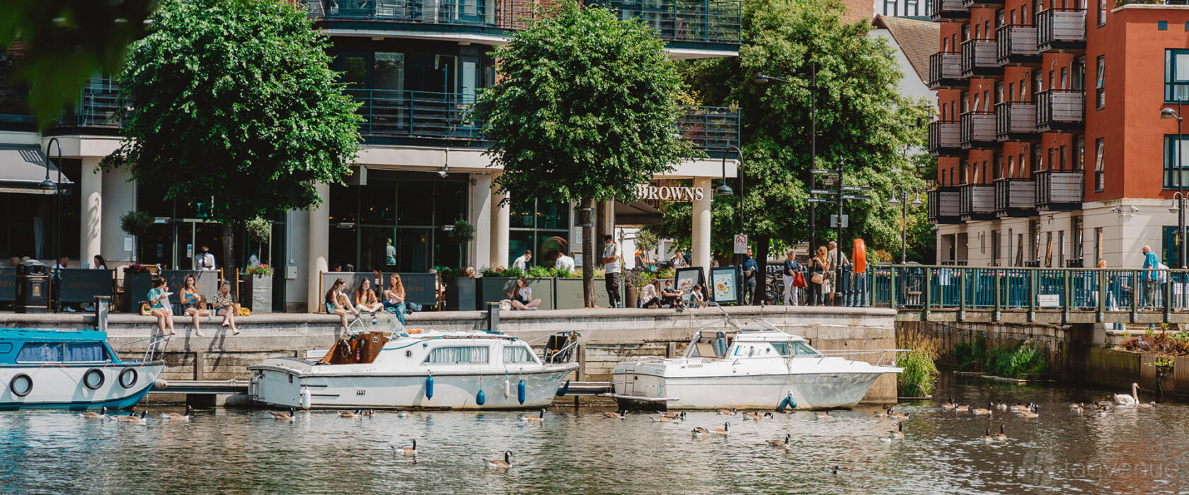 A riverside restaurant with outdoor seating, glass facade, and boats docked along the water at Browns Kingston.
