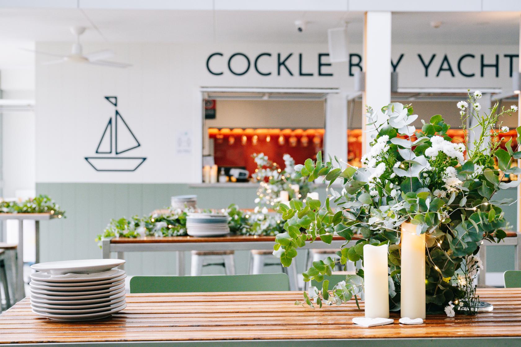 A bar with wooden tables adorned with greenery, candles, and stacked plates at Cockle Bay Yacht Club.