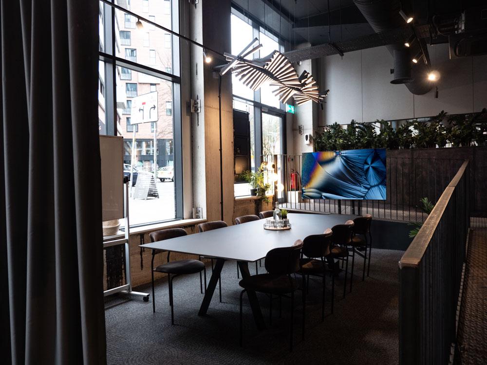 A meeting room with floor-to-ceiling windows, a long conference table, modern lighting, and plants at Colony Piccadilly.