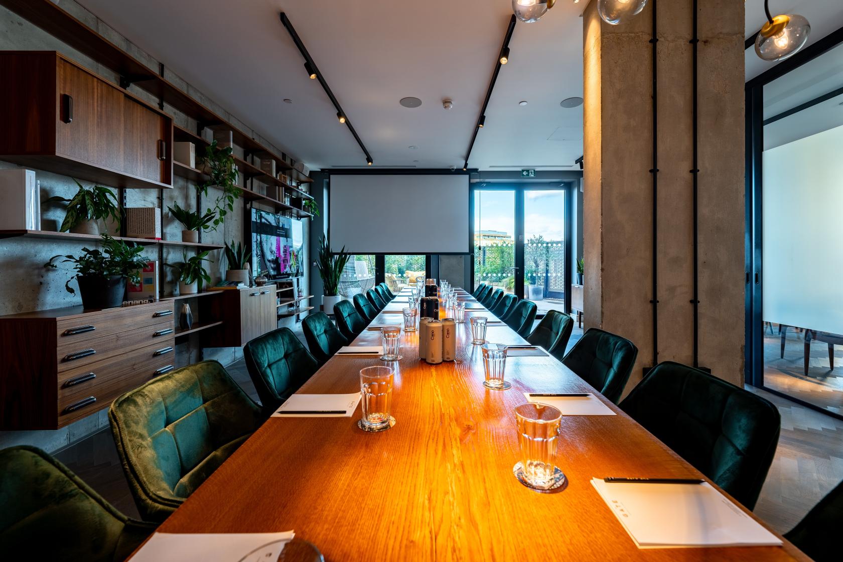 A meeting room with a long wooden table, green chairs, notebooks, glassware, and floor-to-ceiling windows at The Gate London City.