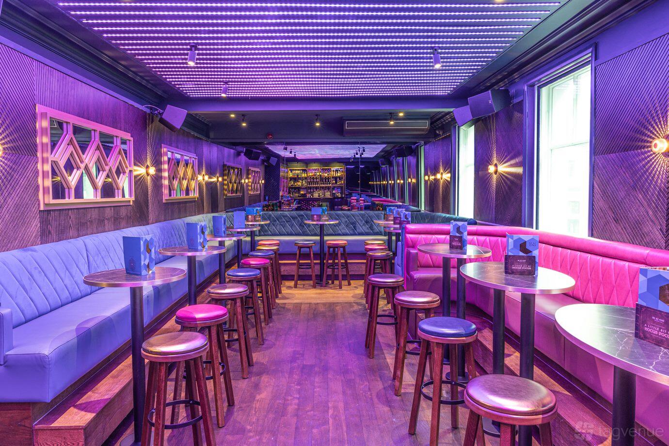 An event space in a bar with purple LED ceiling lights, plush banquette seating, and round high tables at Be At One Leeds Greek Street.
