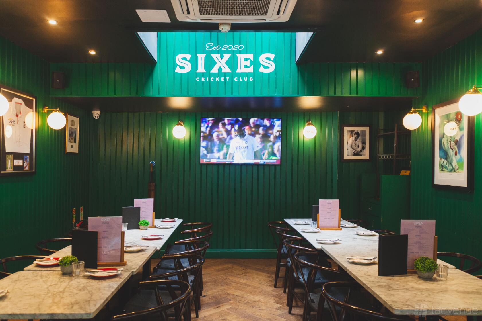 A bar with green paneled walls, sports memorabilia, marble tables, and a large TV at Sixes Social Cricket - Fulham.