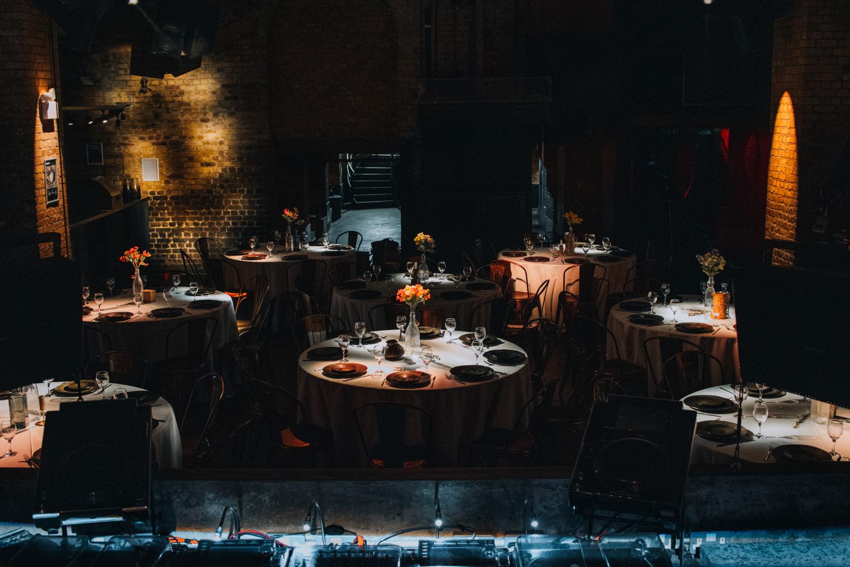 An event venue with round tables draped in white linens, brick walls, and dim ambient lighting at fabric industrial arts events venue.